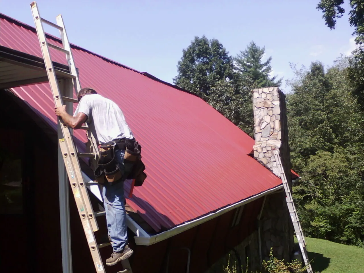 Roofer on a ladder beside a red metal roof and stone chimney — Soffit & Fascia Repair by Roofing Brothers in Towanda